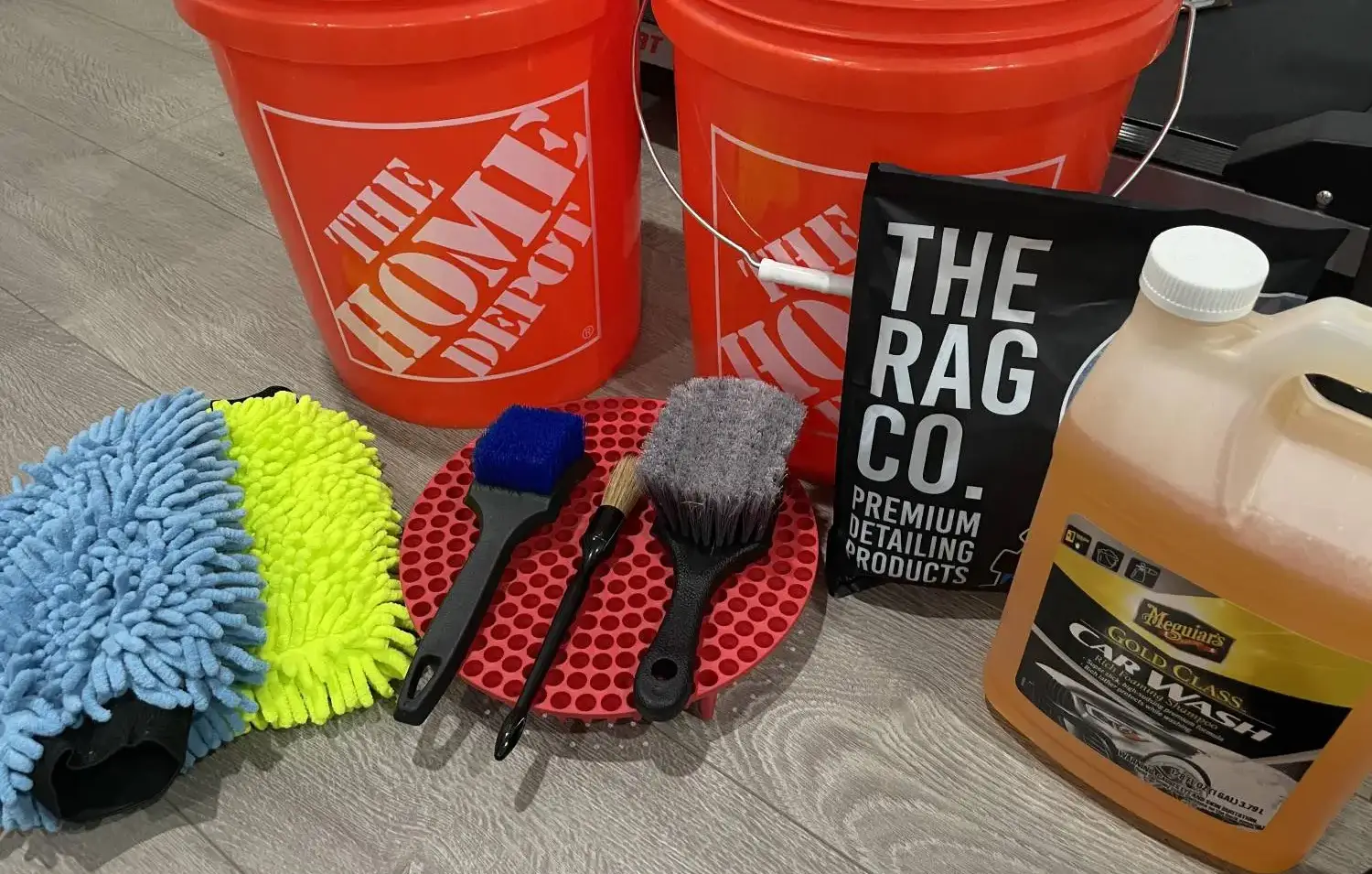 Two-bucket wash setup with grit guards, wash mitts, detailing brushes and a gallon of car wash shampoo.