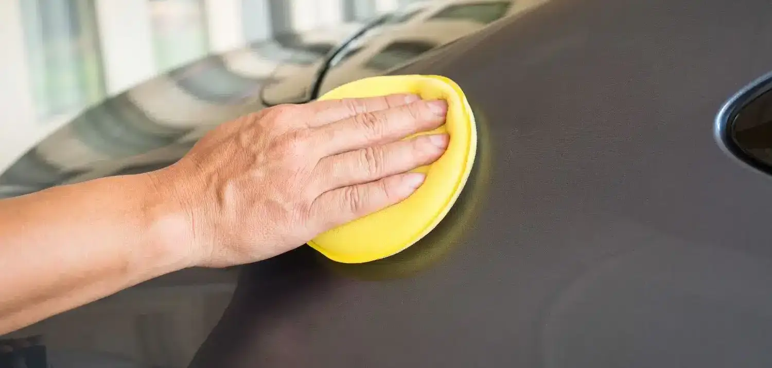 Hand applying car wax with a yellow applicator pad on vehicle surface