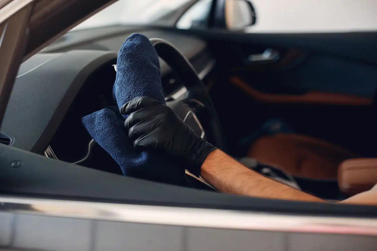 Gloved hand wiping a steering wheel with a blue microfiber towel inside a tan leather interior