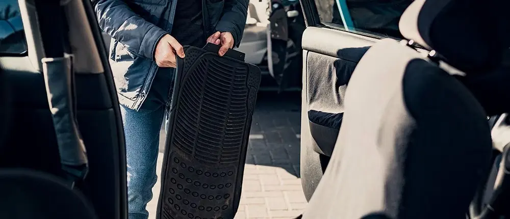Person removing black rubber floor mat from car interior for cleaning.